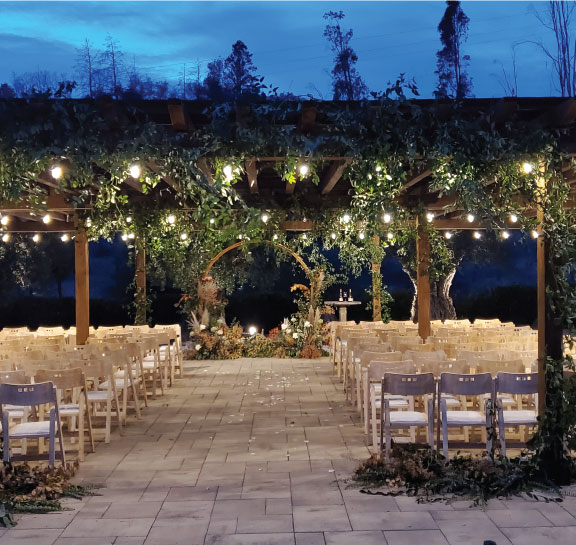 Gorgeous Outdoor wedding venue at Chateau St. Jean featuring white chairs, a wooden canopy, green trees with lights, and a beautiful focal piece for ceremonies