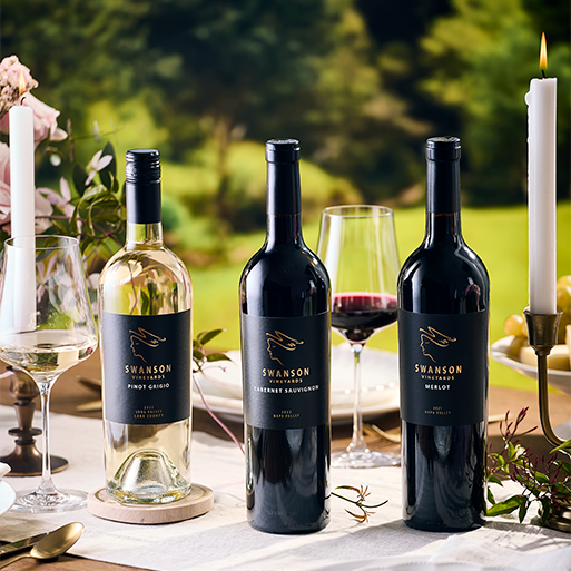 Swanson wines on an outdoor table with candles, place settings, and a poured glass of red wine with trees in the background