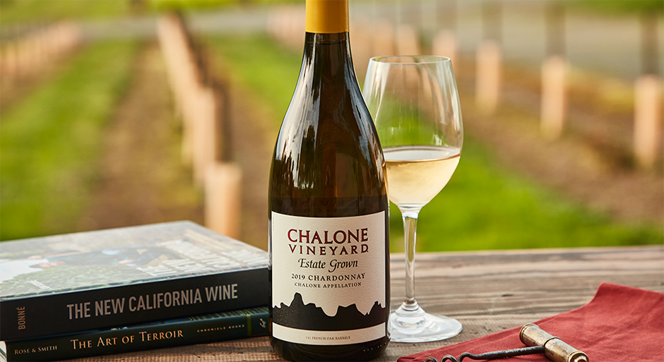 A bottle of chalone wine on a table next to a glass of Chalone white wine, a book, and a view of the vineyard in the background
