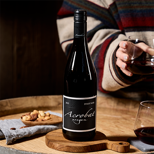 A bottle of Acrobat Pinot Noir on a table with a bowl of nuts and a person holding a glass of wine