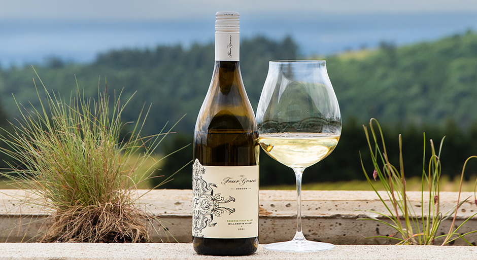 a bottle of four graces wine next to a glass of white wine with the vineyard in the background