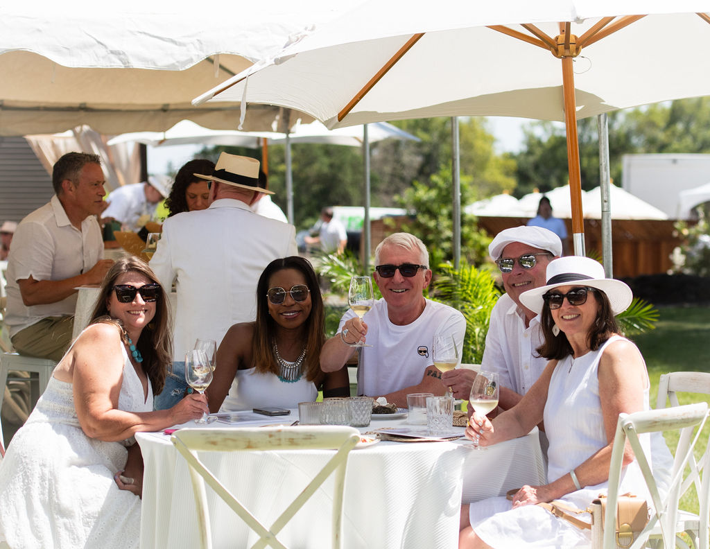 A group of people dressed in summer whites sitting around a table at the party drinking white wine
