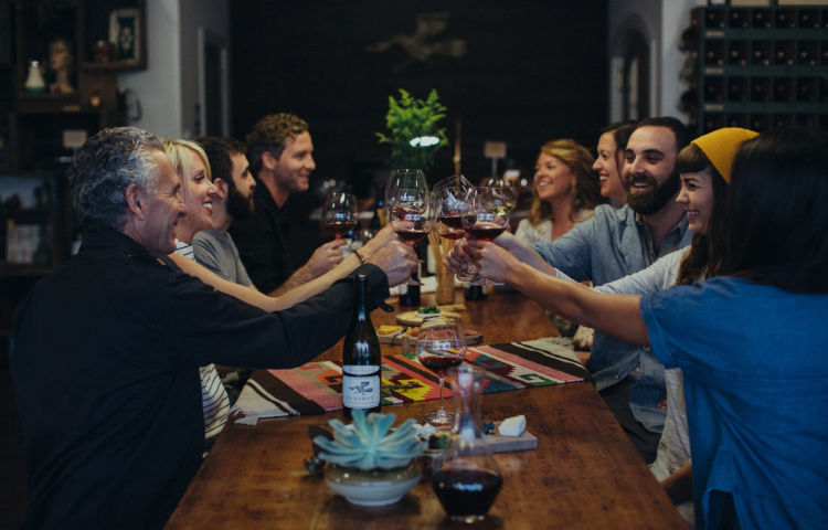 A large group of people sitting around a table, smiling and toasting with glasses of red wine