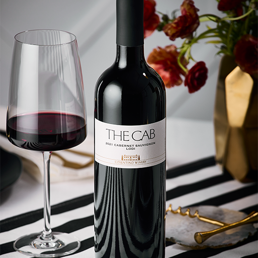A bottle of Cosentino THE CAB wine on a black and white stripped table cloth with flowers in the background and a poured glass of the red wine