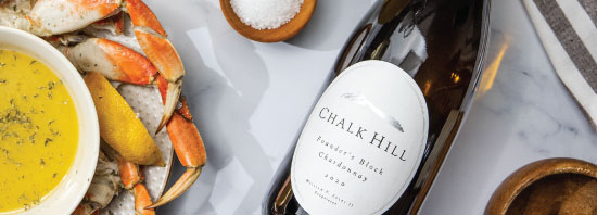 Chalk Hill white wine bottle on a table next to a delicious plate of crab, bowl of soup, and fresh lemons