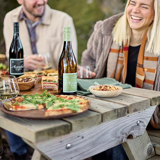 A picnic table setting with pizza and acrobat wine and two people laughing