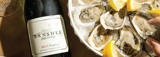 Banshee Ten of Cups Sparkling Wine bottle lying next to a plate of fresh oysters with lemons and a person's hand reaching for an oyster.