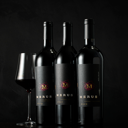 Three vintages of Merus wine with a glass of red wine on a dark, moody background