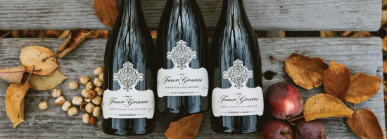 Bottle shot of three Four Graces wine bottles against a wooden backdrop, surrounded by vibrant fall leaves, autumn colors, hazelnuts, and assorted fruits