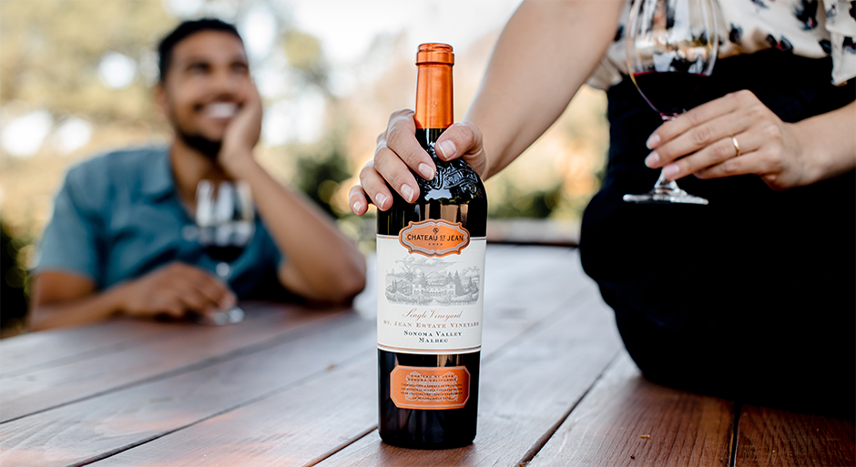 A person with their hand on a bottle of Chateau St Jean wine next to a glass of the wine on a table and a person smiling in the background